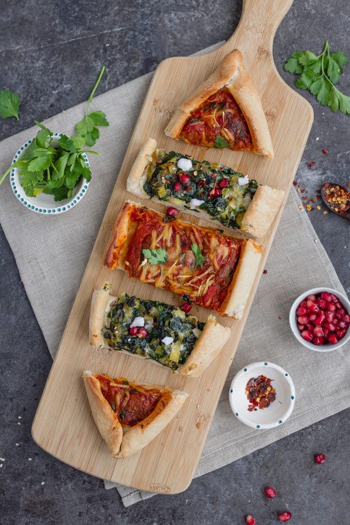 combination of two vegan pide including spinach, leek and vegan feta plus aubergine, tomato and red peper. Toppings in small bowl include pomegranate, chilli flaked and parsley